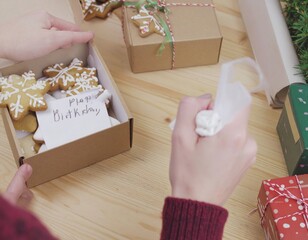 Hands Decorating Snowflake Shaped Cookies With White Icing In A Cardboard Box Surrounded By Gift Wrapped Presents On A Wooden Tabletop