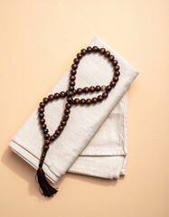 A String of Dark Wooden Prayer Beads With a Tassel Rests on a Folded Light Gray Fabric Square Against a Soft Peach Background