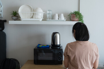Back view of a young woman standing in a modern studio apartment kitchenette with a microwave, electric kettle, and organized shelving