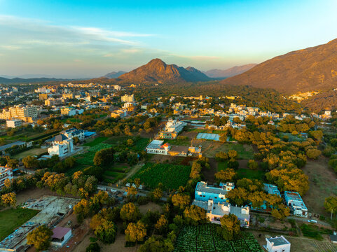 Aerial view of a small town nestled among lush greenery, with mountains looming in the background, casting long shadows in the golden light, Pushkar Rural, Rajasthan, India.