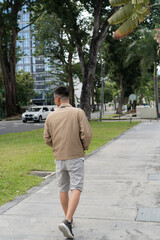 Fototapeta premium Rear view of a young Asian man in a beige jacket walking along a tree-lined city sidewalk with a modern high-rise building in the background