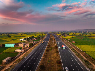 Aerial view of a modern highway cutting through vibrant fields under a dramatic sunset sky, connecting distant horizons, Alwar, Rajasthan, India.