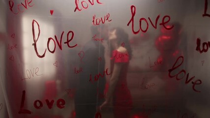Zoom in view of transparent polyethylene film with word love written multiple times with red paint in foreground, and young couple holding hands and kissing on lips on Valentines day in background