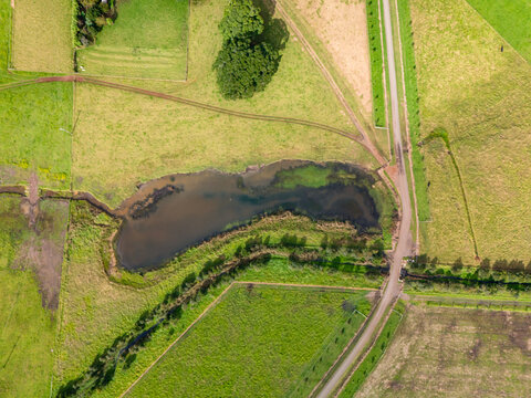 Aerial view of a serene pond surrounded by vibrant green fields and a narrow road cutting through, Crooked River Road, Gerroa, New South Wales, Australia.