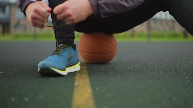 White athlete ties blue running shoe kneeling on asphalt court by yellow line, basketball placed behind. Hands deftly loop laces while jacket sleeves push back, closeup lowangle view capturing