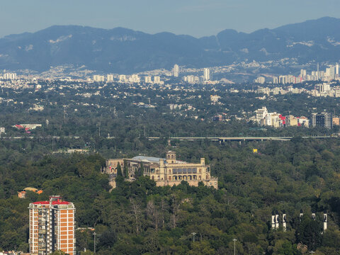 Aerial view of Chapultepec Castle crowning a verdant hill amidst a sprawling cityscape painted in muted tones, framed by distant mountains, Mexico City, Mexico.