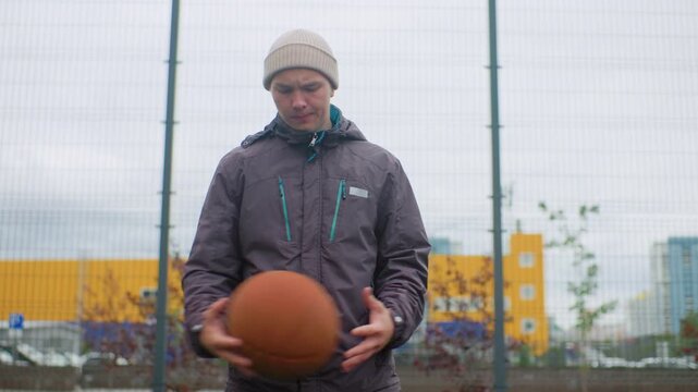 white man with basketball on court wearing knit beanie and waterproof jacket dribbling with steady rhythm under cloudy sky hoop and chainlink fence frame scene highrise apartments in background