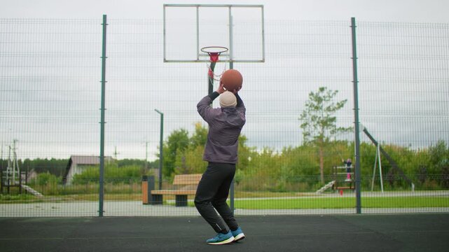 senior white man shoots basketball outdoors at fenced suburban court, performs jump for rim dunk wearing jacket and sneakers, focused expression during solitary training session under cloudy sky,