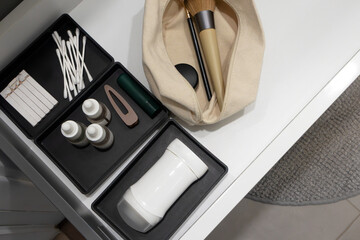 Modern bathroom drawer with neatly organized skincare bottles, cotton swabs, hair accessories, deodorant and makeup brushes in fabric pouch, top view