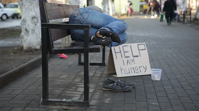 A dirty homeless man in torn socks lies on a bench in a city park and asks for alms. The inscription on the cardboard, Help, I am hungry.
