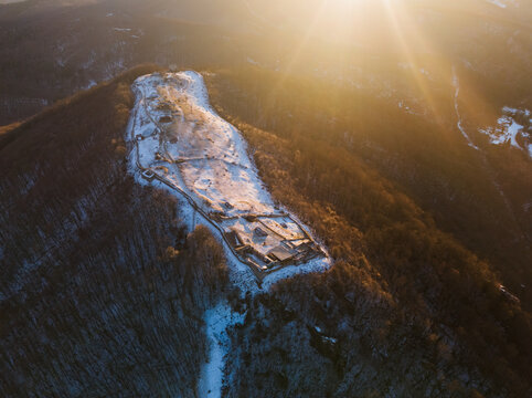 Aerial view of the snow-dusted Pusty hrad ruins rise majestically against a golden sunset, casting long shadows on the forested slopes, Zvolen, Banskobystrick&Atilde;&frac12; kraj, Slovakia.