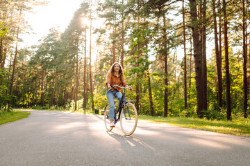 Naklejka premium A young woman rides her bike along a sunny park path. A happy woman enjoys a bike ride through the trees. Concept of nature, weekends, and relaxation.
