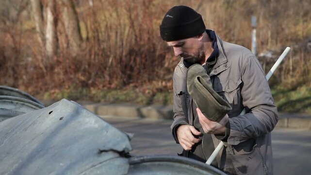 A dirty male homeless man is looking for shoes in a dumpster. A homeless person rummages through garbage bins.