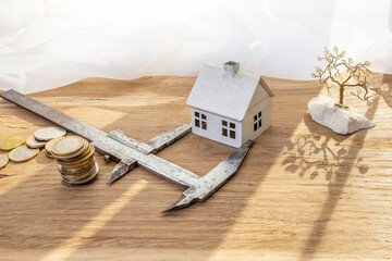 Metal caliper measuring a white model house next to a stack of coins on a wooden desk, construction cost assessment concept