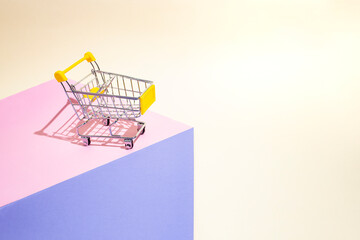 Pop art style photography of a shopping cart on purple , pink and beige paper background arranged as a geometric podium, abstract consumerism metaphor