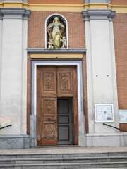 Fototapeta premium St. Mary of the Assumption parish church facade in Calendasco, Emilia Romagna, Piacenza, Italy, featuring a wooden entrance door, stone steps, and a statue of the Virgin Mary Assumption with cherubs