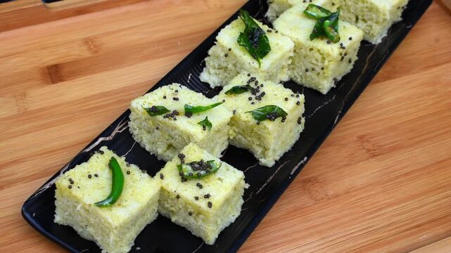 light green dhokla served on a black platter, garnished with green chilli, mustard seeds or rai, and curry leaves. 