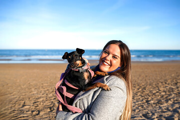 Happy woman holding her small black dog on the beach and smiling at camera