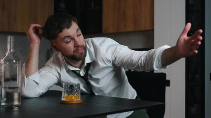 Adult man in a white shirt sitting alone in a dark room and drinking strong alcohol. Empty and half-empty bottles and a glass on the table in front of him. Concept of addiction alcoholism