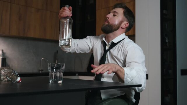 A very drunk alcoholic man sits and dances in a dark kitchen after drinking strong alcohol.