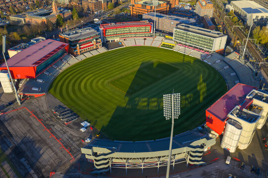 Aerial image of the Old Trafford Cricket Ground in Manchester, featuring the green pitch, red hospitality wings, and the long shadow of a floodlight tower during the golden hour.