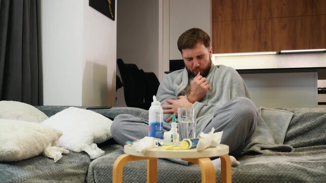 Sick man suffering from a cold wrapped in blanket sitting on sofa in living room at home. A tired bearded male coughs, sneezes, blows his nose with a handkerchief. He has fever or flu.