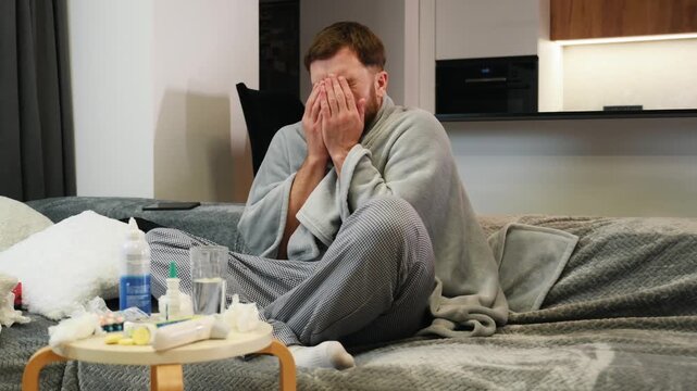 Sick man suffering from a cold wrapped in blanket sitting on sofa in living room at home. A tired bearded male coughs, sneezes, blows his nose with a handkerchief. He has fever or flu.