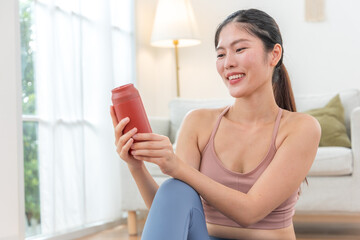 Smiling Asian woman resting after home workout with water bottle.