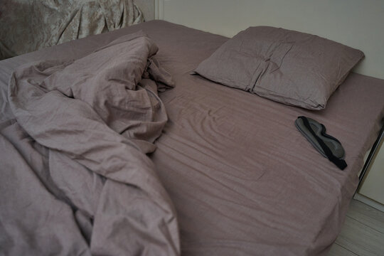 Unmade mauve bed with sleep mask on pillow in modern cozy bedroom setting.