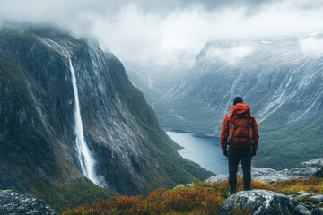 Obraz premium A lone hiker stands on rocky terrain, admiring a majestic waterfall cascading down steep cliffs into a serene fjord below. The misty atmosphere enhances the natural beauty of the landscape.