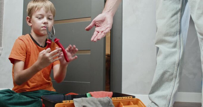Curious boy sitting on floor pulls toolbox closer and hands pliers to father during calm home repair process. Cozy domestic atmosphere highlights learning trust teamwork