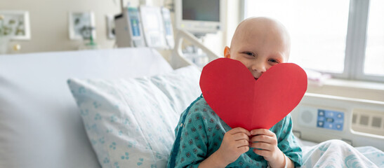 Bald Caucasion Child with red paper heart in hospital bed. Concept of health, hope, recovery for Valentine's Day or childhood cancer with copy space.