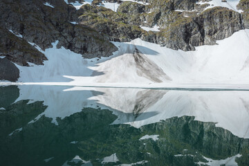 Ideal horizontal symmetrical snowy image in reflection of glacier lake. Smooth mirror pure water surface with snowfield. Stony rocky wall with glacier above alpine lake. Snow symmetry in mountains.