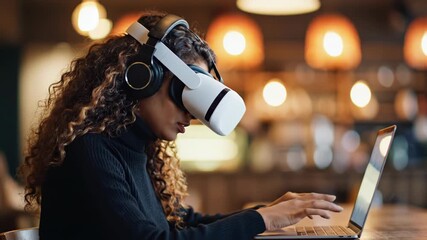 Woman wearing virtual reality headset and headphones while using laptop in a modern coffee shop with warm lighting