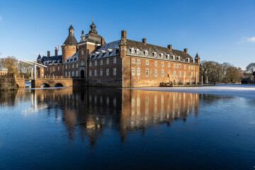 Fototapeta premium Majestic brick castle with towers and dormers reflects in partially frozen moat, framed by leafless trees and clear winter sky.