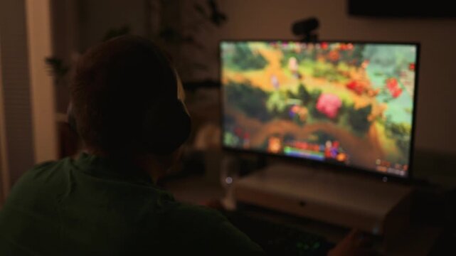 Caucasian teenager wearing headphones plays online video game on PC, spends leisure time at home. Professional young gamer enjoys gameplay in 3D third person shooter on computer. Gaming and Esports