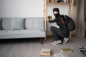 Female thief in balaclava looking at lens during a house robbery. Ransacking room for cash. burglary, property theft, and illegal break-in. Criminal woman searching for loot.