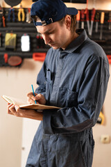Professional male carpenter standing in his woodworking workshop and writing notes in notebook
