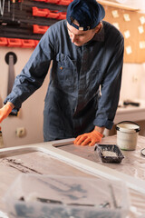 Skilled male worker in protective workwear painting wooden furniture in professional carpentry workshop
