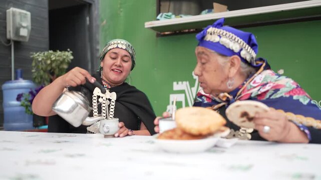 Two indigenous women in traditional clothing enjoying sopaipillas and mate tea at an outdoor table