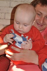 Baby dressed in red intently examines their sock-covered foot while being held by an adult. The child's focused expression highlights their curiosity and exploration.