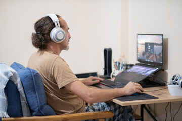 Focused Gamer Using Laptop and Headset. Man Playing Video Games on Laptop with Headphones. Home Gaming Lifestyle with Laptop. Online Gaming at Home with Laptop.