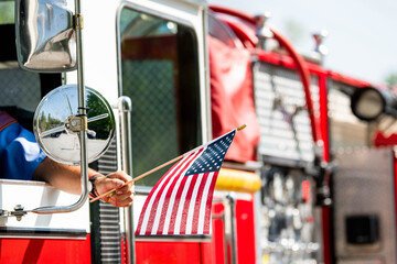 A firefighter waves an American flag from the driver's seat window of a firetruck during a patriotic US parade for Memorial Day, Flag Day, or July 4th. © Jeanne Sager Photo