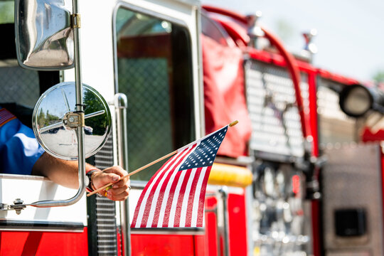 A firefighter waves an American flag from the driver's seat window of a firetruck during a patriotic US parade for Memorial Day, Flag Day, or July 4th.