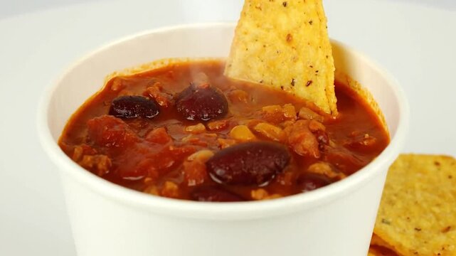 A cup of steaming chili with a single tortilla chip dipping in and chips in a white surrounding