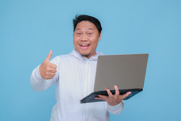 closeup of a young adult man raising his thumb in front of his laptop while laughing broadly like an advertisement wearing white t-shirt on turquoise blue  