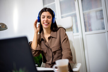 Young woman enjoying music on headphones in her home office. Cozy workspace with laptop and coffee, ideal for relaxation, work-from-home, and lifestyle concepts.