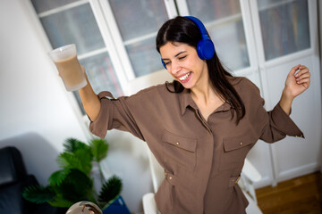 Young woman playing on her laptop while holding a cup of coffee and listening to music on headphones. Cozy home lifestyle, digital leisure, and multitasking enjoyment.