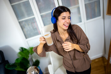 Young woman playing on her laptop while holding a cup of coffee and listening to music on headphones. Cozy home lifestyle, digital leisure, and multitasking enjoyment.