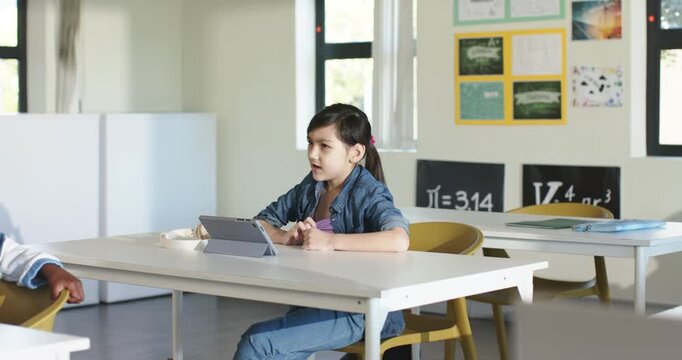 Asian child turning to classmate, returning to tablet tapping doing work at desk in denim jacket